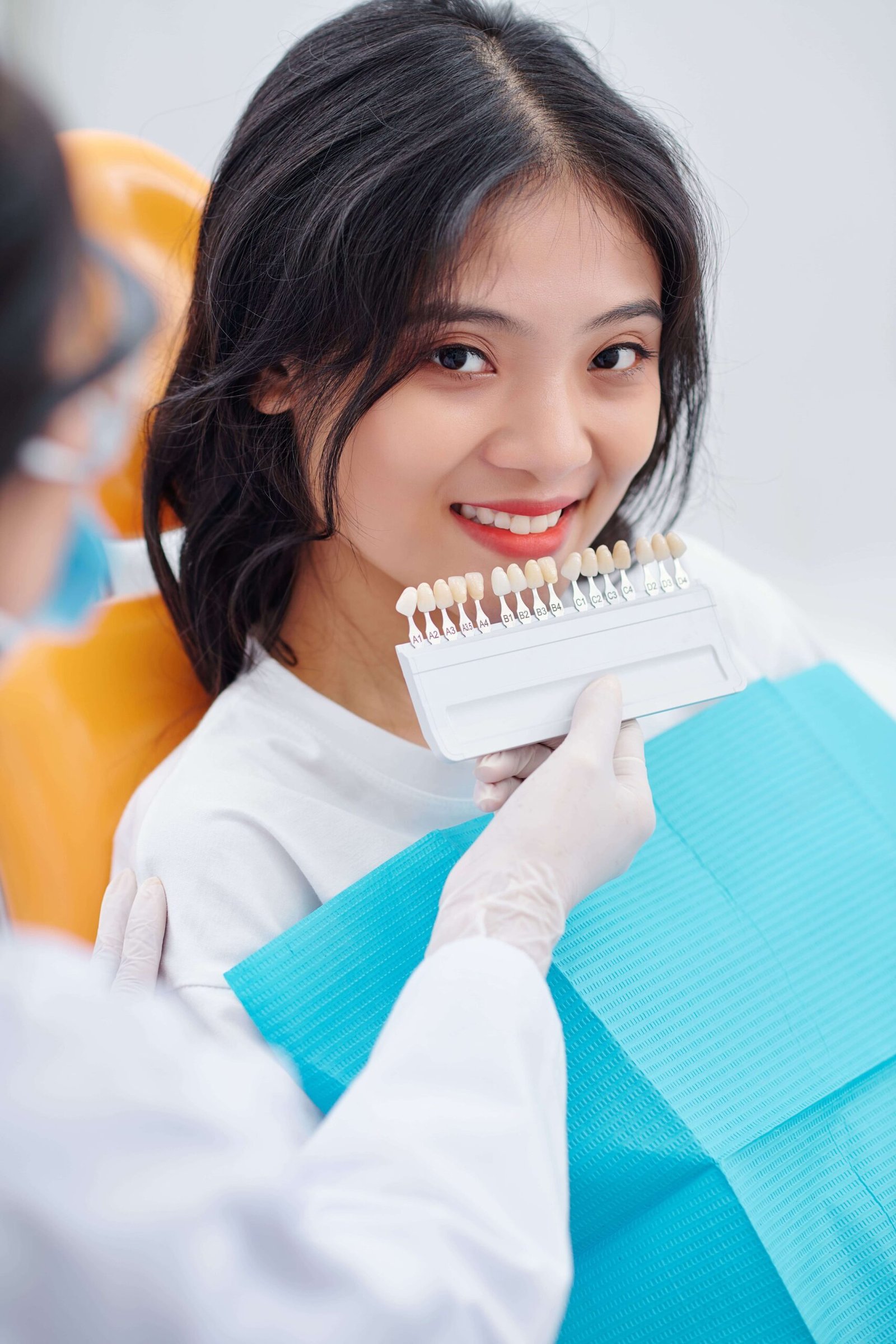 A dental professional, wearing a lab coat and white gloves, holds a shade guide up to a smiling female patient. The patient has dark hair and a bright, confident expression as she looks at the camera, symbolizing the trusted and personalized process for Dental Veneers in Ajman.